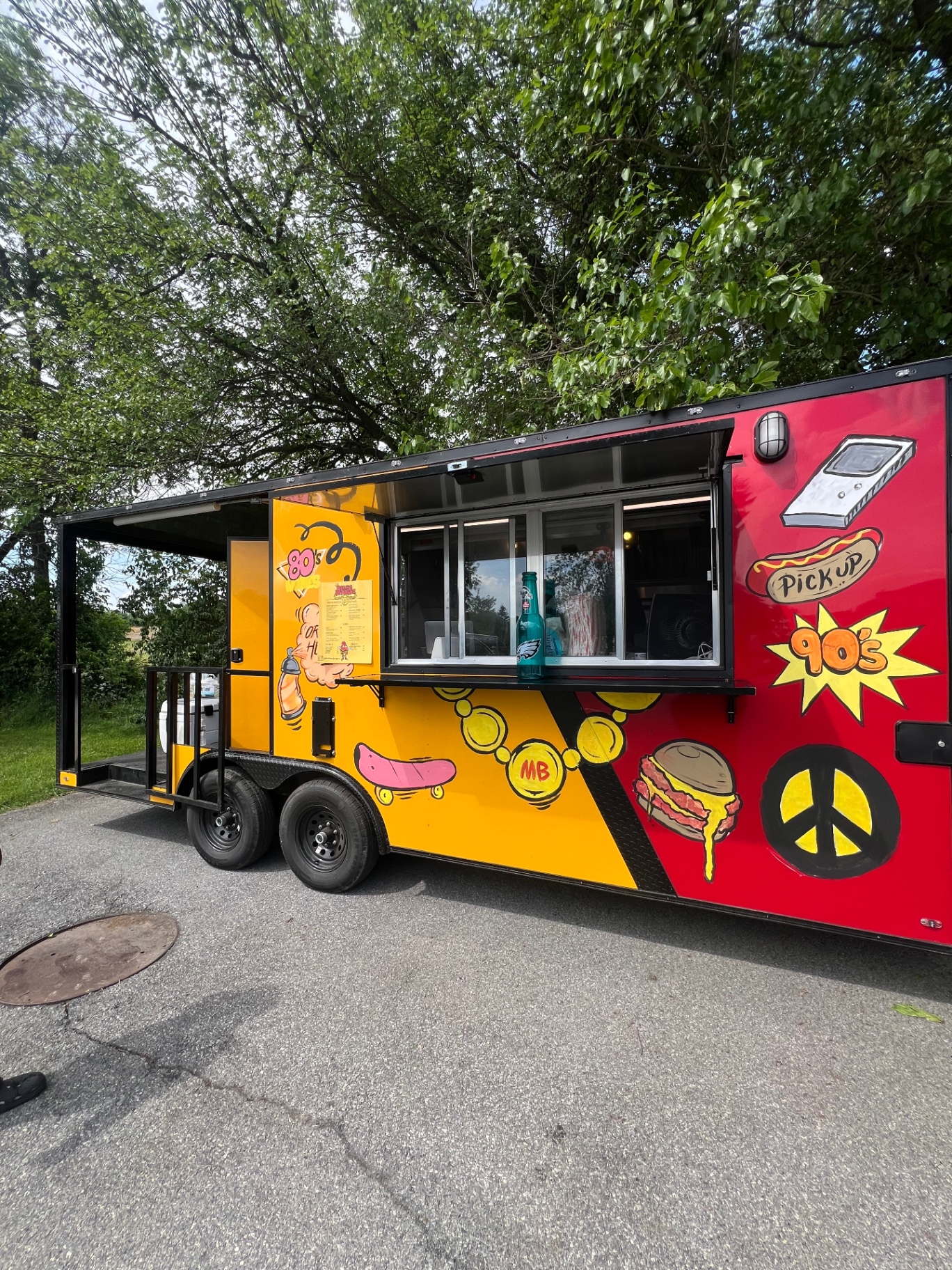 Side view of the red and yellow Mallie's Burgers food truck with 80s and 90s graffiti art, pickup window open under the trees