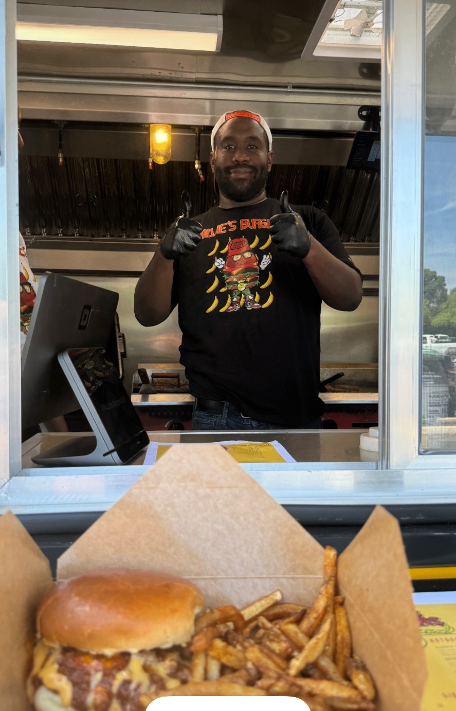 Mallie's Burgers cook in a branded tee giving two thumbs up from the food truck service window above a chili cheeseburger and fries