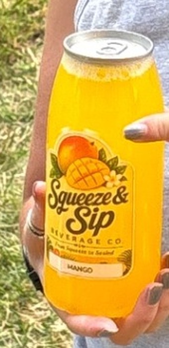 Close-up of the Squeeze & Sip mango lemonade label on a can held by a smiling customer at a Mallie's Burgers car show setup